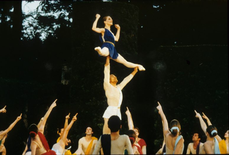 A dancer lifts another dancer on stage surrounded by a dance company striking a pose below them