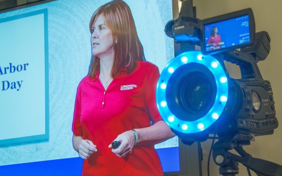 A person in a red shirt on a tv screen next to a studio camera.