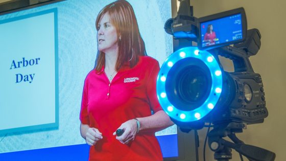 A person in a red shirt on a tv screen next to a studio camera.