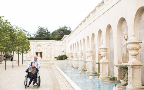 someone steering another in a wheelchair next to a stone fountain 