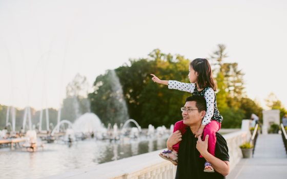 Kids sitting on shoulders of an adult pointing at fountains