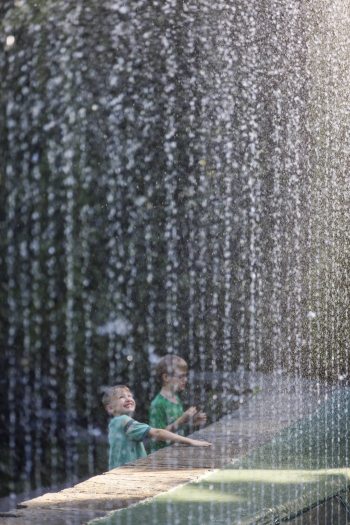Through a curtain of small water streams, a small child reaches their hand into the water