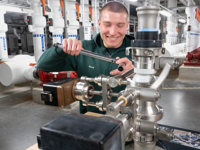 A person holding a wrench smiles as they work on a piece of machinery in a pumproom