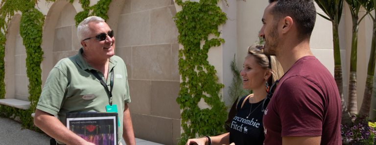 A Longwood staff member holding a binder of information speaks to a couple in in front of a vine covered wall