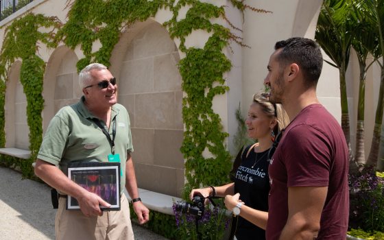 A Longwood staff member holding a binder of information speaks to a couple in in front of a vine covered wall