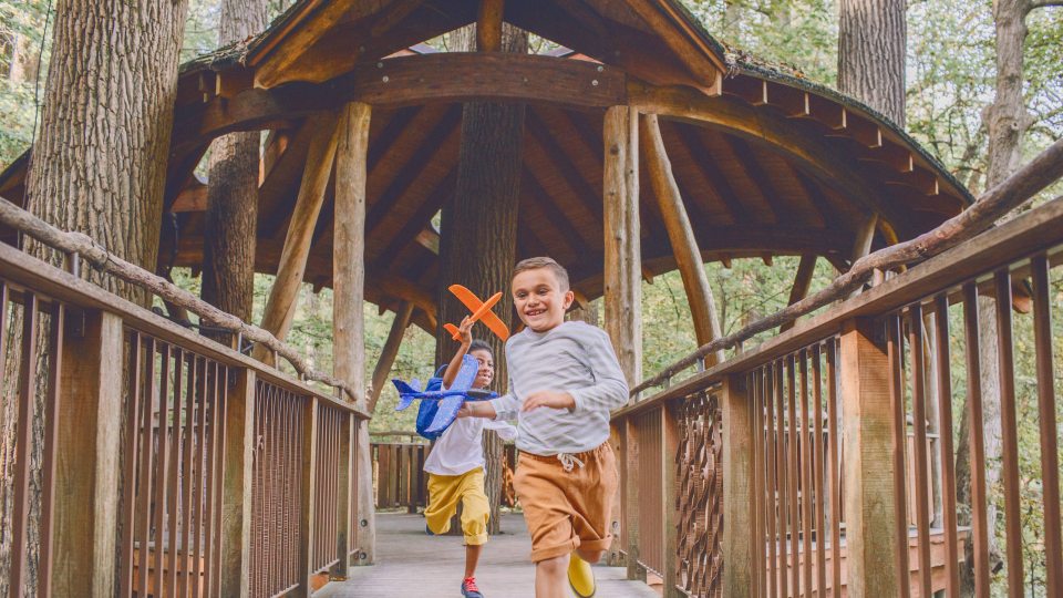 Two children holding toy airplanes run along a wooden walkway out of a wooden treehouse