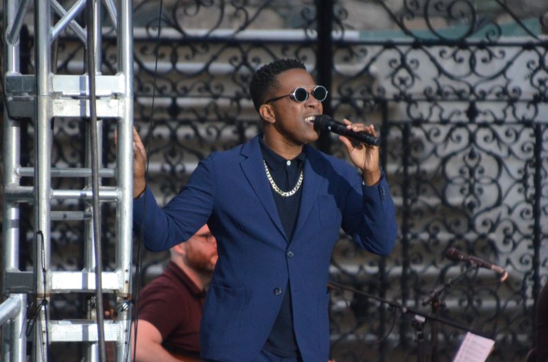 A man in a blue suit sings on stage into a handheld microphone