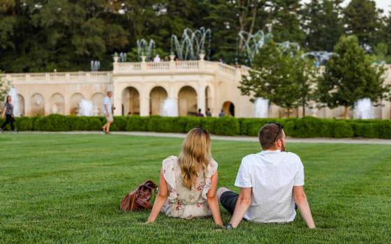 Two people relax on a green lawn while fountains dance in the background.