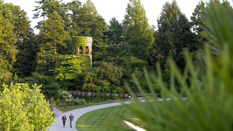 Two guests walk along a curved garden path amid lush greenery and a stone carillon tower in the background.