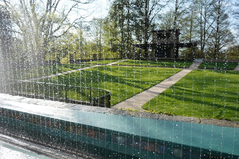 Fountains of tiny streams of water shoot up from a stage in front of a green grass seating area