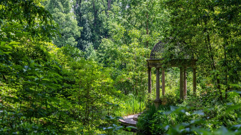 A stone gazebo sits beside a small lake tucked into a forest of bright green trees