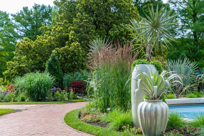 ornamental grasses, bold tropical foliage, and pale green containers near a brick pathway and blue pool of water
