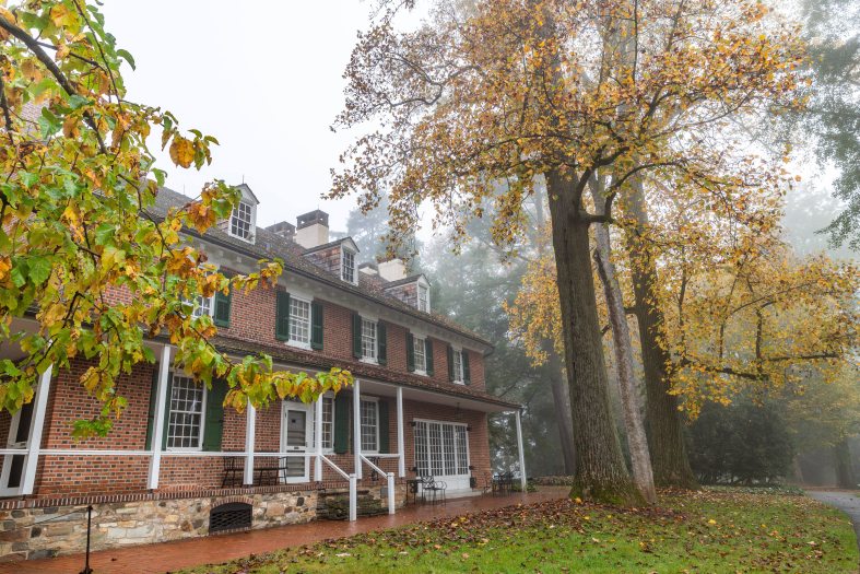A stone and brick house is seen surrounded by autumn trees of yellow and orange and a morning fog