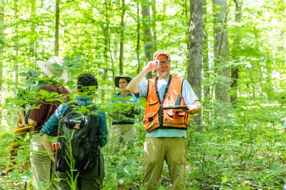 A man in an orange safety vest holds up a botanical sample in a lush green forest with three onlookers. 