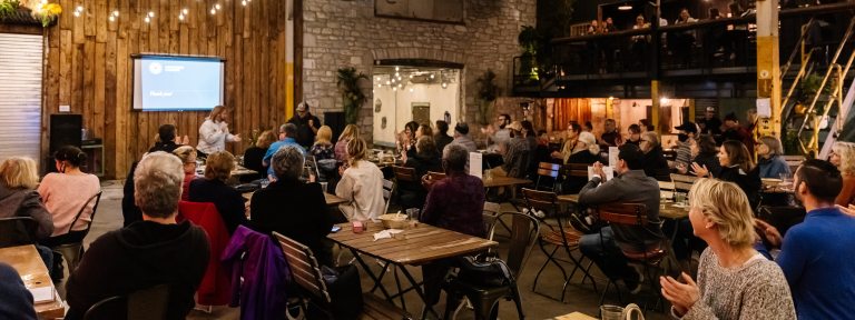 people sitting at tables in a pub-like atmosphere clap for a speaker giving a presentation