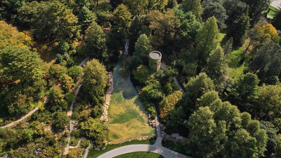 Aerial view of large grassy area with a small pond and stone tower surrounded by a forest