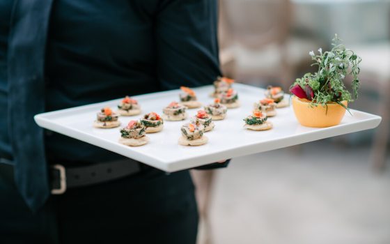 Closeup of server dressed in black, holding a white tray of appetizers, with an edible arrangement decorating one corner of the tray.