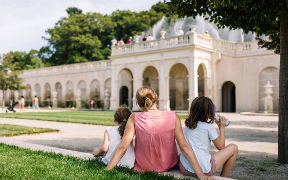 An adult and two children rest on a white curb at lawn's edge, facing a facade of fountains.