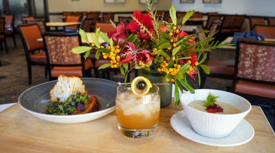 a plate of food, drink, bowl of soup and a vase of flowers on a wood table