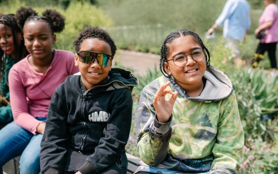 Students stitting on a stone wall in a garden smiling at the camera.