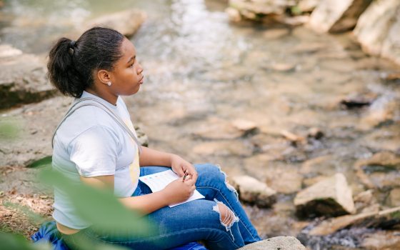 A person sitting on a large rock next to a pond writing in a notebook.