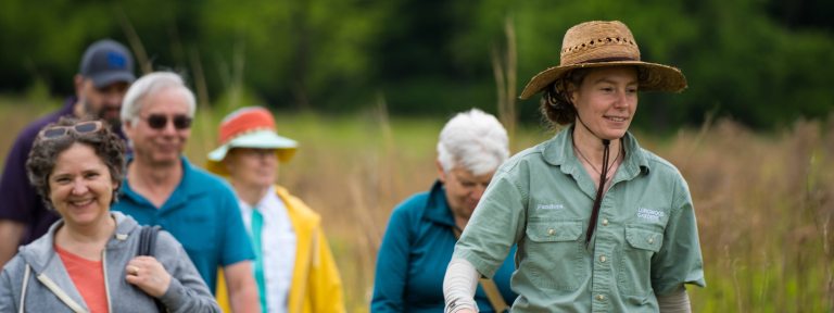 Smiling people follow a guide in a hat on a walk through a meadow.