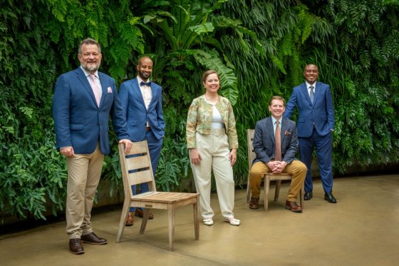A group of five people posed in front of a green wall smiling at the camera.