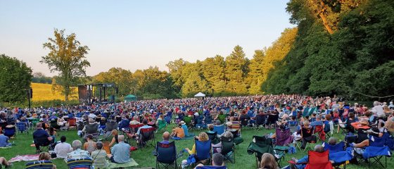 An outdoor concert overlooking a meadow with people sitting in chairs looking at the stage.