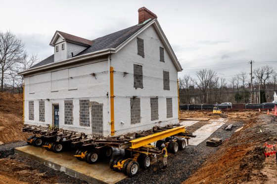 An old white house being moved on a large trailer with wheels.
