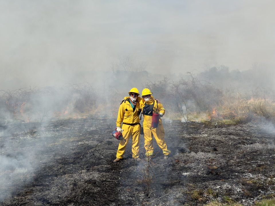 Two people in yellow protective suits standing in a recently burned meadow.