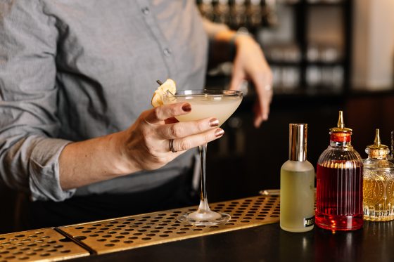 A person's hands behind a bar holding a cocktail glass preparing to hand it to someone. 