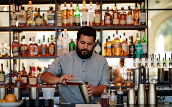 A bartender pours a drink at a bar that is plentifully stocked with a variety of liquor on shelves in the background.