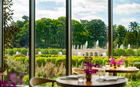 A view from a restaurant table looking out through large windows. In the background, a grand stone fountain structure with multiple water jets is surrounded by lush green trees and manicured hedges under a bright, partly cloudy sky.