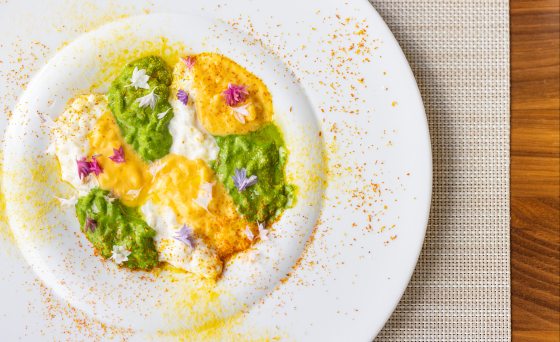 Close-up, overhead view of a plated dish, likely risotto, with alternating dollops of creamy white, vibrant green, and yellow puree or sauce, garnished with edible flowers and surrounded by spice dust.