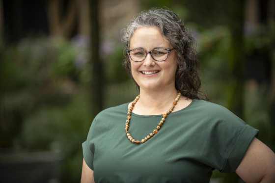 Close-up smiling headshot of a woman with gray, curly hair, glasses, a green shirt, and a beaded amber necklace.