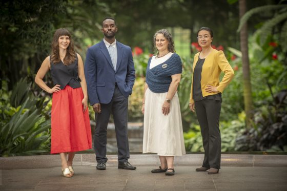 A full-body group portrait of four Longwood Fellows standing side-by-side in formal business-casual attire against a lush green background.