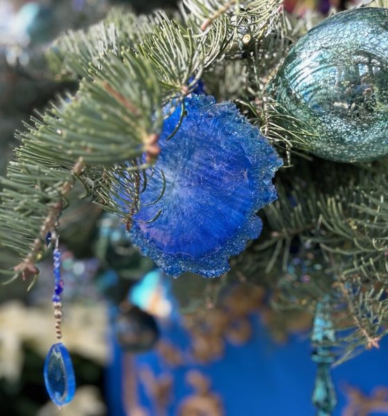 A close-up shot of a Christmas tree branch adorned with blue and turquoise ornaments. A striking, fuzzy, royal blue floral or star-shaped ornament is visible, along with a hanging decoration featuring a dangling, tear-drop-shaped clear blue crystal bead. White poinsettias are visible and slightly blurred in the lower left background