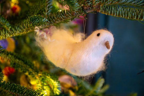 A close-up of a Christmas tree branch with a fluffy, white felted fiber art ornament shaped like a small bird hanging from it.