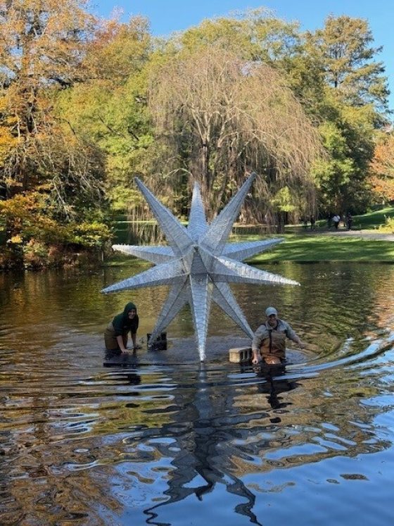 Two workers installing a large, silver, spiky star-shaped Christmas light display in a small lake, with autumn foliage visible around the water.