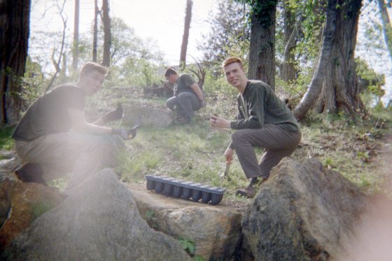 Students Micah High, Matthew Fleischhacker, and Travis Cox are seen planting on a wooded hillside. They are crouching among rocks and trees, with a tray of starter plants in the foreground.