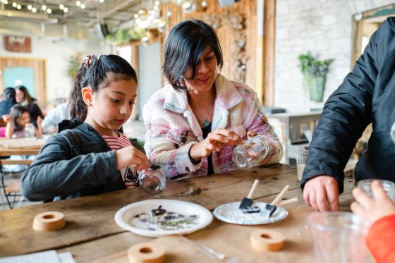 A woman and a young girl sit side-by-side, carefully applying glue and pressed botanical elements to glass jars using foam brushes.