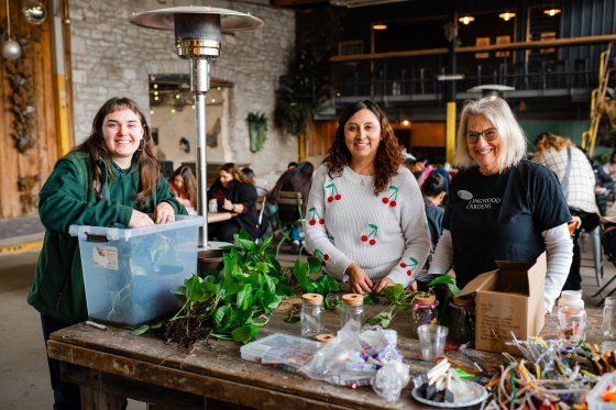 Three smiling women—workshop organizers and staff—stand behind a rustic wooden table filled with supplies including green vines, glass jars, corks, and colorful pipe cleaners.