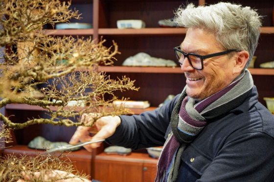 A candid shot of the man in the glasses and scarf smiling as he works on the tree. He is using long, thin bonsai shears to carefully trim the fine outer growth of the Elm.