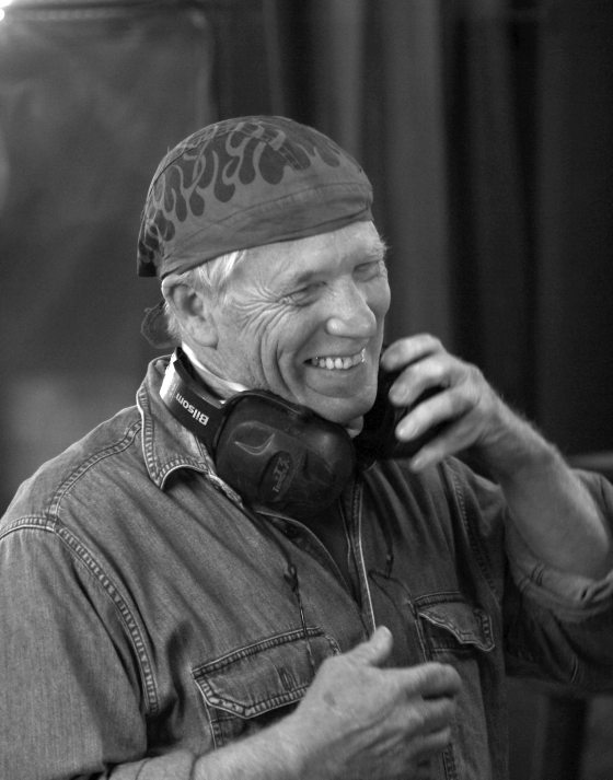 Black & white portrait of person smiling broadly, wearing a headscarf and denim shirt, with headphones around their neck.