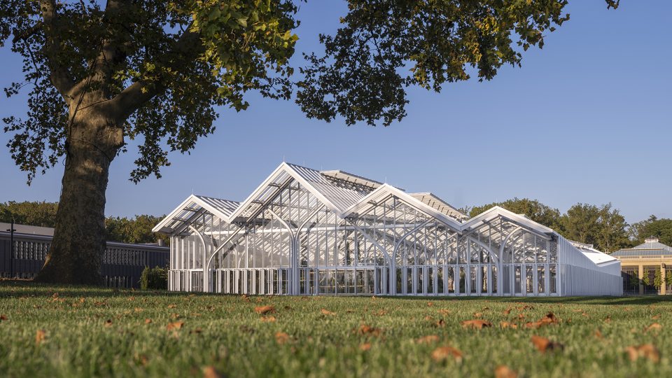 a glasshouse with a multi-peaked roof rises from a wide green lawn dotted with fallen autumn leaves