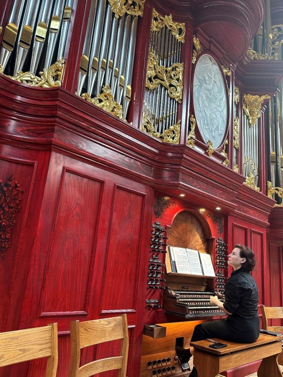 A person seated at a large organ, with silver pipes towering towards the ceiling. 