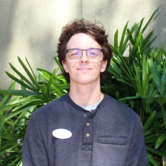 Portrait of person with wavy brown ear-length hair, wearing glasses and a dark blue shirt, head tilted slightly upward.